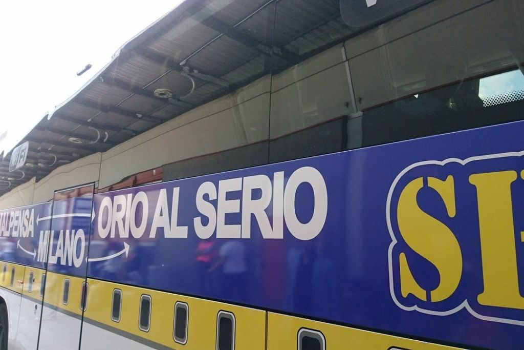 측면에 "ORIO AL SERIO" 라고 쓰여진 파란색과 노란색 버스가 금속 지붕 구조물 아래에 주차되어 있습니다.