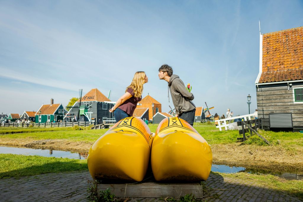 네덜란드 전통 가옥과 풍차를 배경으로 잔디가 우거진 지역에서 두 사람이 커다란 노란 나무 막사 위로 키스를 하고 있습니다.