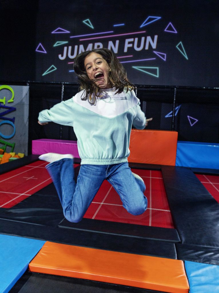 다채로운 색상의 폼 블록과 "JUMP N FUN" 이라고 적힌 표지판이 있는 실내 놀이 공간에서 한 어린이가 트램펄린에서 즐겁게 뛰어놀고 있습니다.