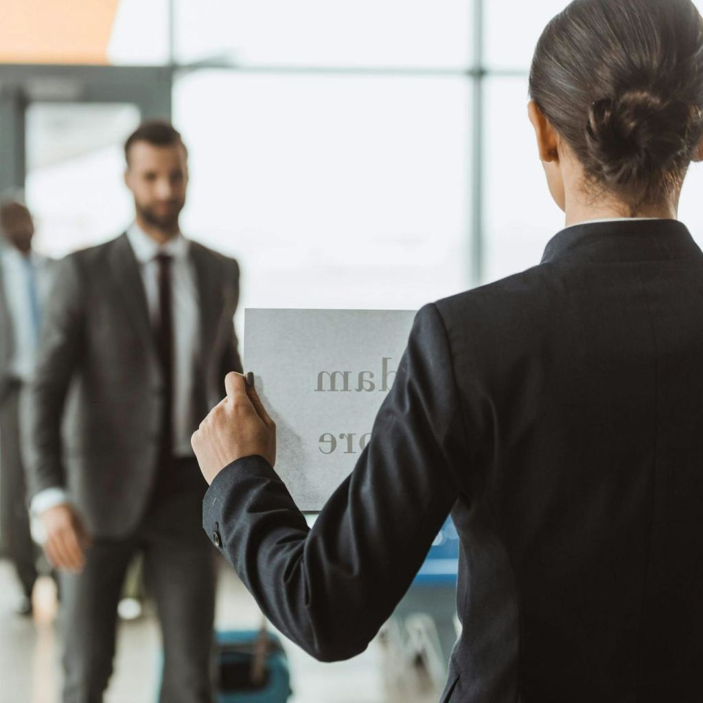 양복을 입은 사람이 허리 높이에서 공항을 향해 걸어가는 회색 양복을 입은 다른 사람과 마주하는 표지판을 들고 있습니다.