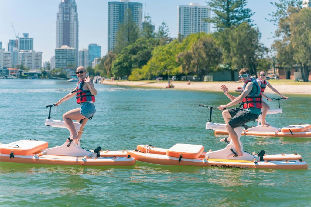도시의 스카이라인과 울창한 녹지를 배경으로 구명조끼를 입고 몸을 흔들며 수상 자전거를 타는 사람.