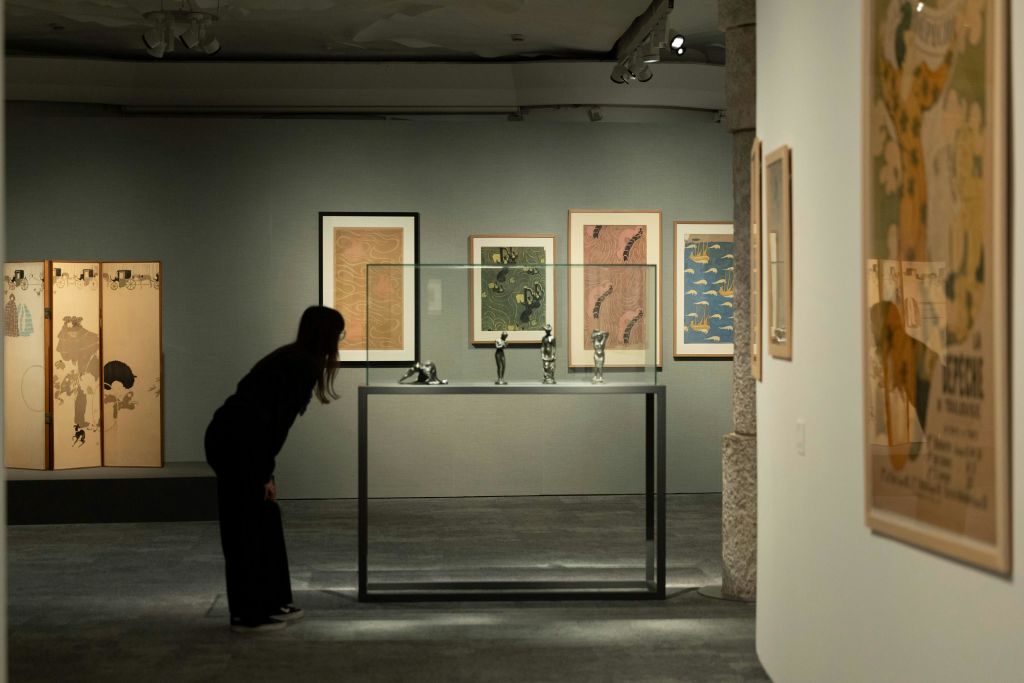 한 사람이 벽에 액자 그림이 그려진 미술관의 유리 케이스에 전시된 작은 조각품을 관찰합니다.