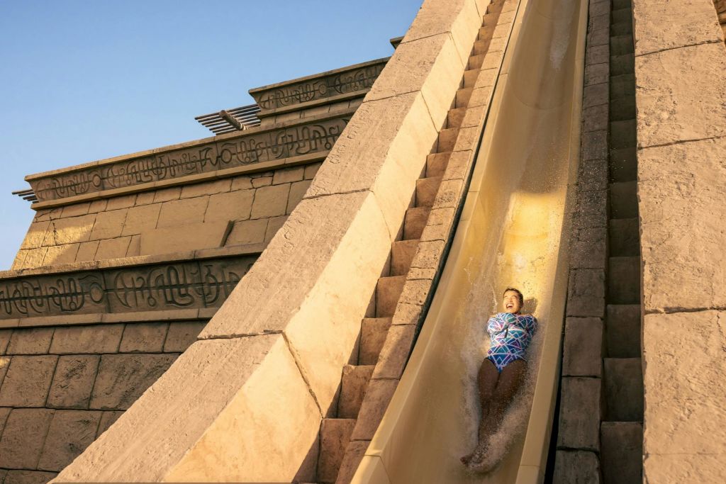 맑은 하늘 아래, 돌과 같은 구조물로 둘러싸인 워터파크에서 가파른 물 미끄럼틀을 타고 내려오는 사람.