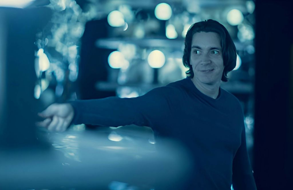 긴 머리카락을 가진 사람이 흐릿하고 둥근 조명을 배경으로 어둡게 빛나는 공간에 서서 미소를 짓고 옆을 바라보고 있습니다.