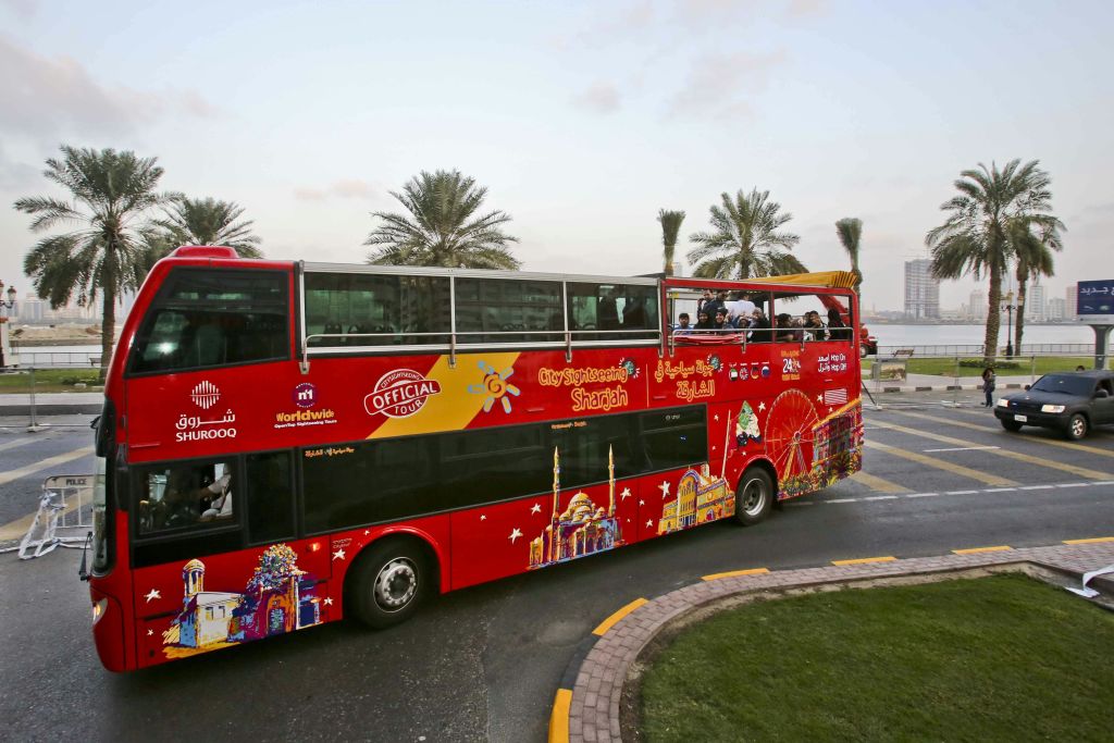 "City Sightseeing Sharjah" 브랜드가 새겨진 빨간색 2층 투어 버스입니다. 탑승객이 오픈탑 상부 갑판에 있습니다. 야자수를 배경으로.