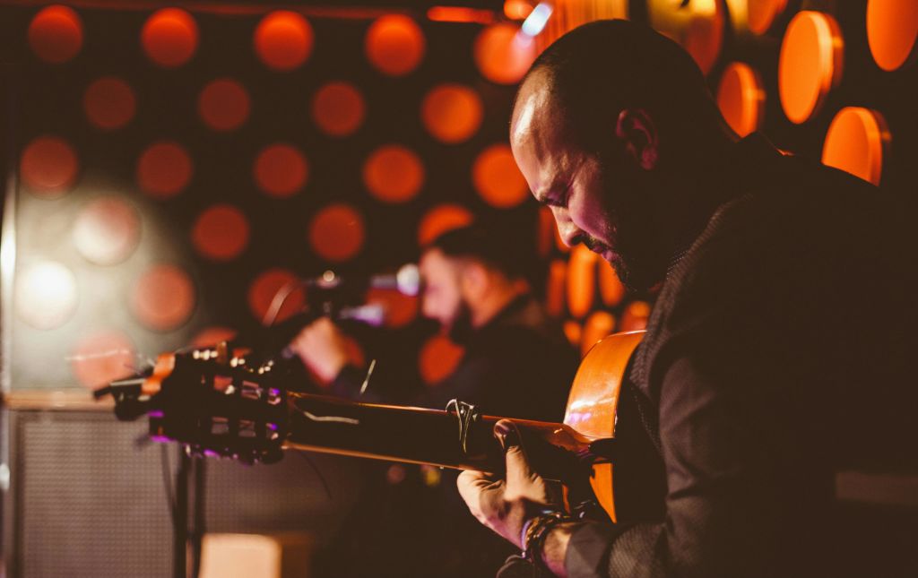 벽에 주황색 원형 조명이 켜진 어두운 방에서 한 남성이 기타를 강렬하게 연주하고 있습니다. 또 다른 뮤지션이 배경이 흐릿해집니다.