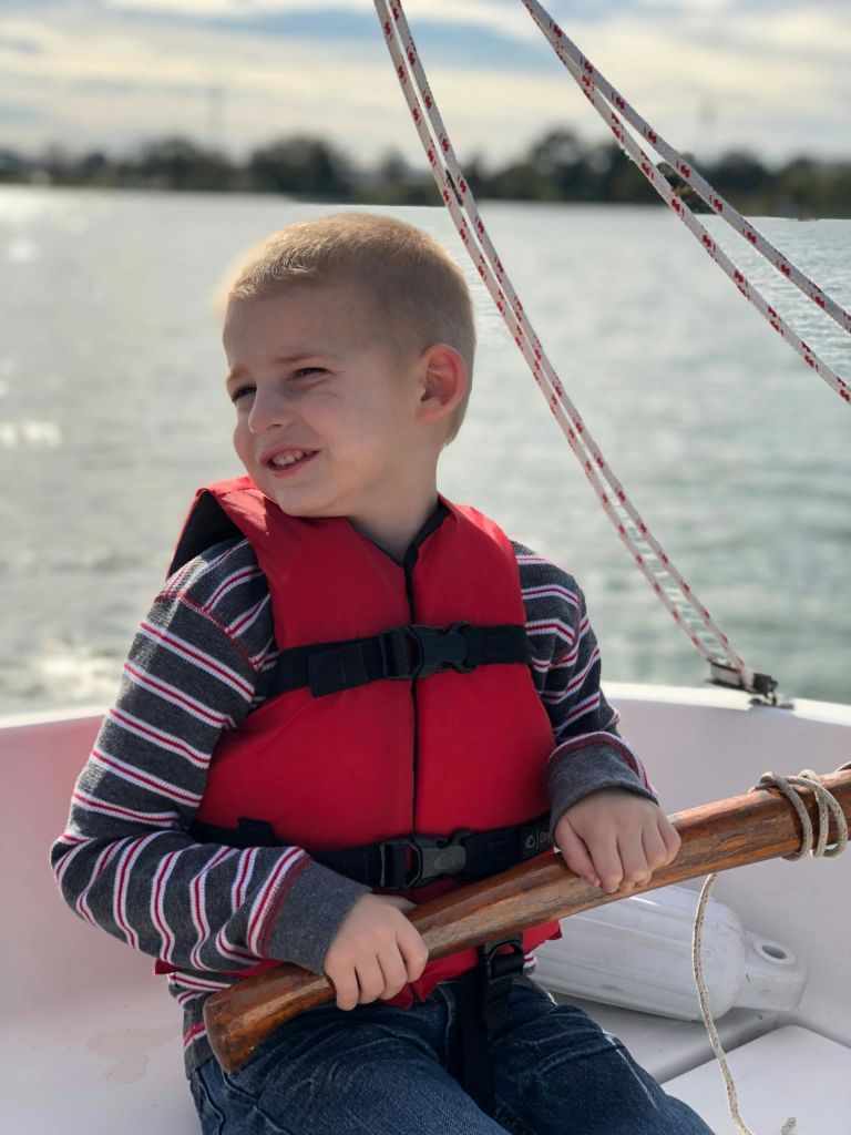 빨간 구명조끼와 줄무늬 셔츠를 입은 어린이가 보트에 앉아 물과 나무를 배경으로 미소를 짓고 있습니다.