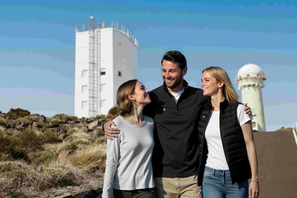 푸른 하늘을 배경으로 하얀 건물 근처 야외에서 미소를 짓고 포옹하는 세 사람, 두 명의 여성과 한 명의 남성.