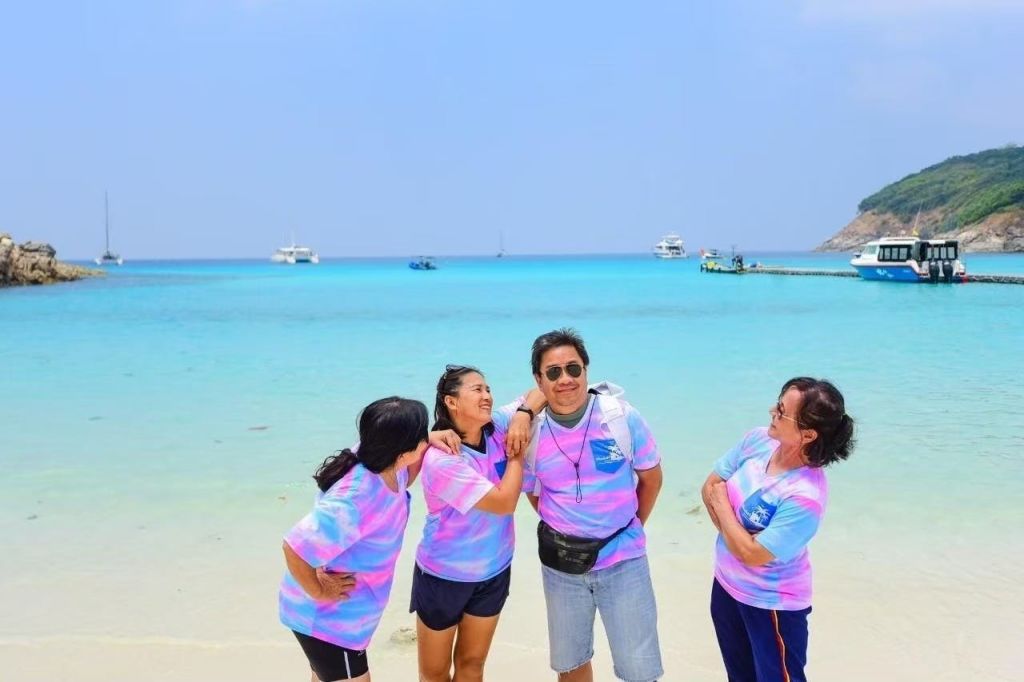 타이 다이 셔츠를 입은 네 명이 맑고 푸른 물과 보트를 배경으로 해변에서 미소를 짓고 포즈를 취하고 있습니다.