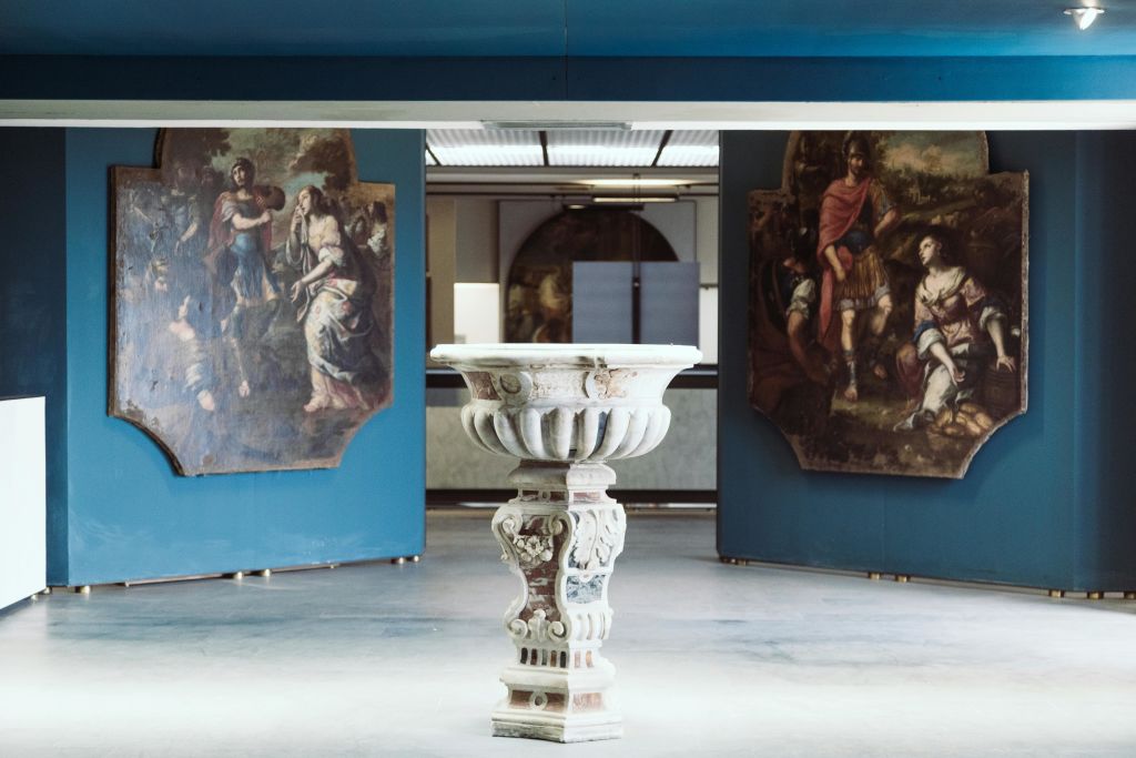 갤러리의 화려한 석조 받침대는 파란색 벽에 있는 커다란 고전 그림과 나란히 놓여 있습니다.