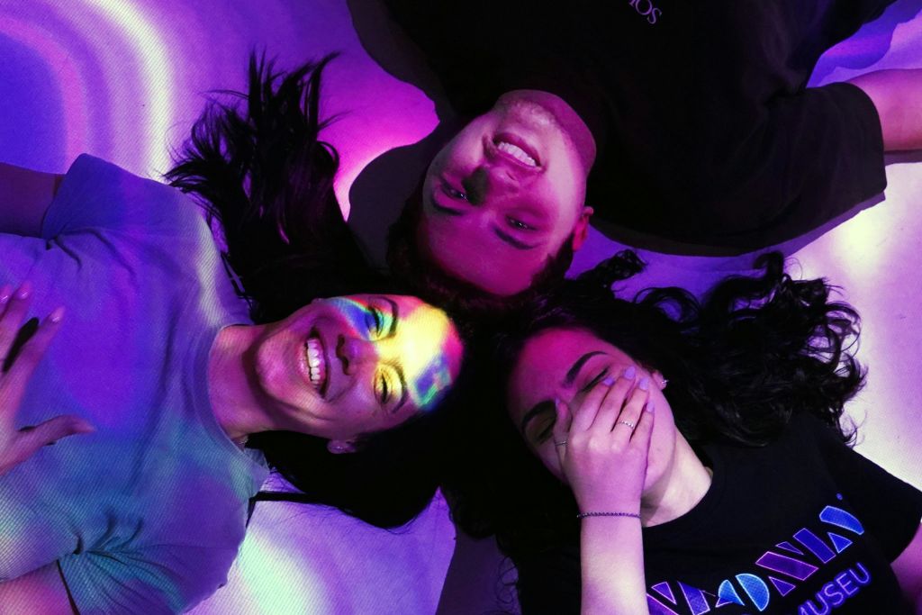 얼굴에 형형색색의 불빛이 투영된 채 등을 대고 누워 웃고 있는 세 사람. 한 사람이 입을 가리고 있다.