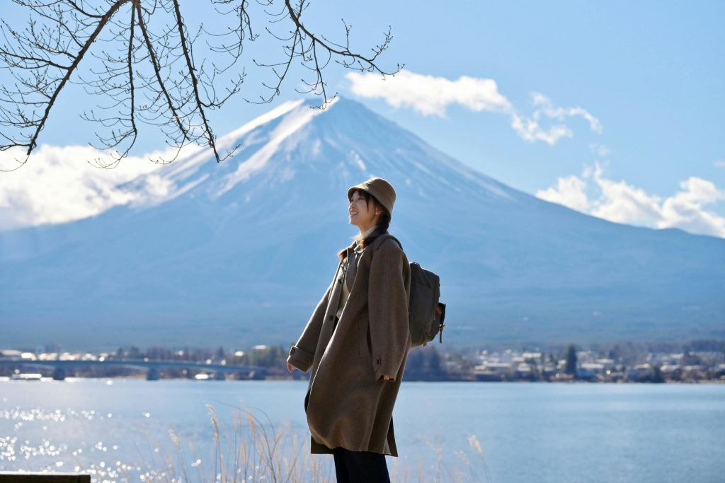 배낭을 메고 코트와 모자를 쓴 사람이 눈 덮인 산을 배경으로 호수 근처에 서 있습니다.