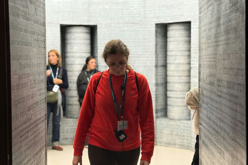 빨간색 스웨터를 입은 여성이 앞쪽에 서 있고, 다른 두 사람이 배경에 서 있으며, 문자로 덮인 벽이 있는 방에 서 있습니다.