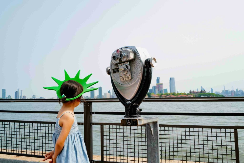 도시의 스카이라인이 내려다보이는 동전으로 작동하는 양안 뷰어 옆에 녹색 자유의 여신상을 입은 어린이.
