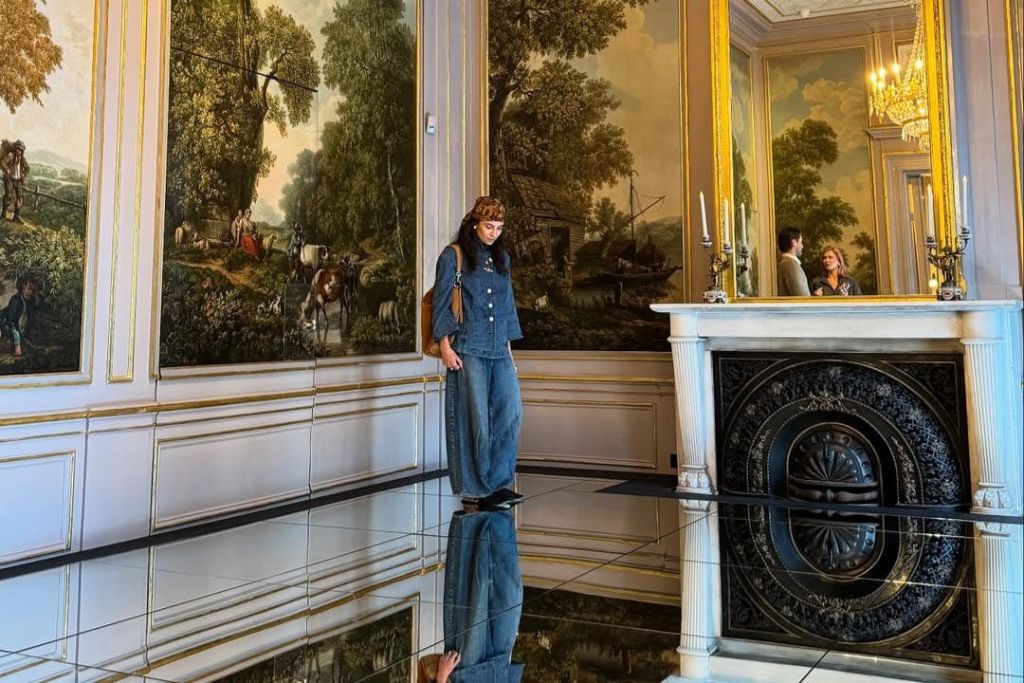 바닥에서 천장까지 이어지는 벽화와 반사 바닥이 있는 화려한 방에 데님 옷을 입은 사람이 서 있습니다.