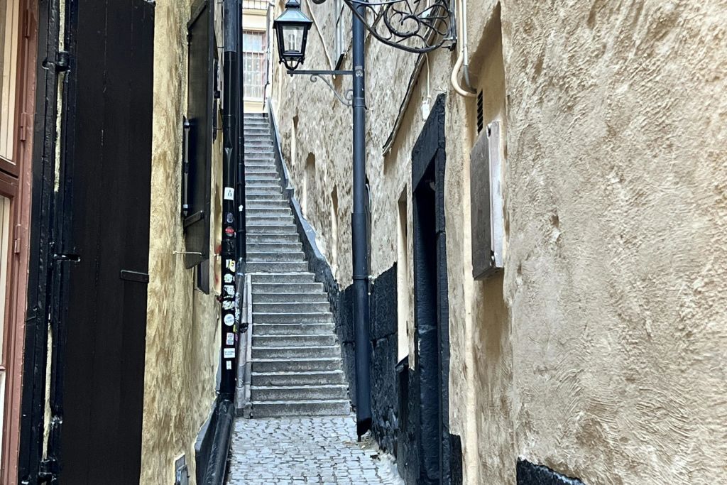 낡고 질감이 있는 건물 사이로 올라가는 가파른 계단이 있는 좁고 자갈이 깔린 골목으로, 벽면 램프가 장식되어 있습니다.