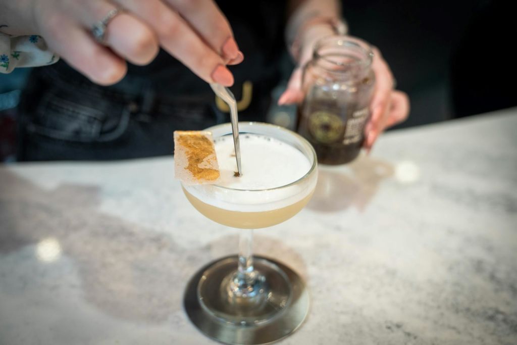 작은 도구로 줄기 유리에 거품이 나는 칵테일을 장식하고 다른 손에 항아리를 들고 있는 사람.