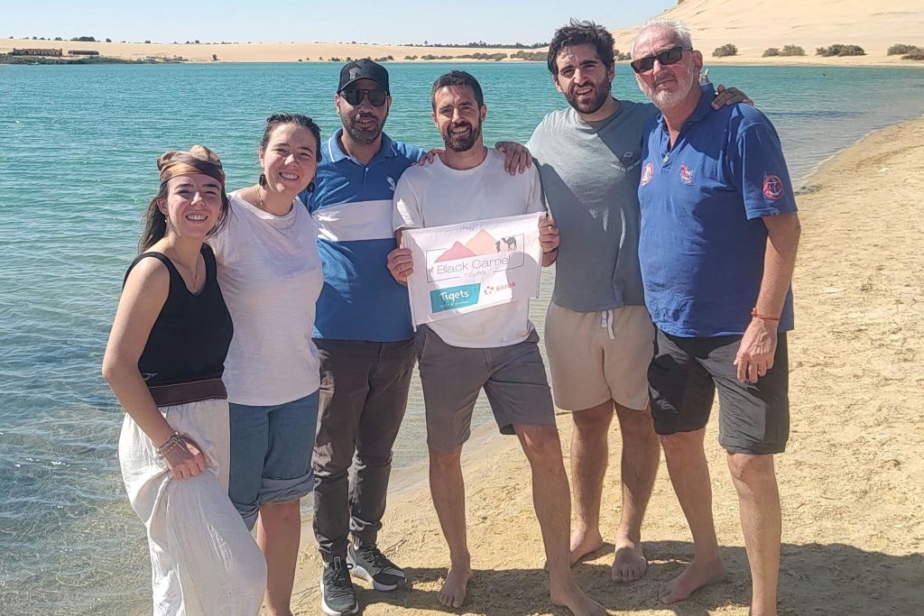 여섯 명이 호숫가의 모래 해변에 함께 서 있으며, 두 명은 "검은 캠프 티게츠" 라고 쓰인 팻말을 들고 있습니다.