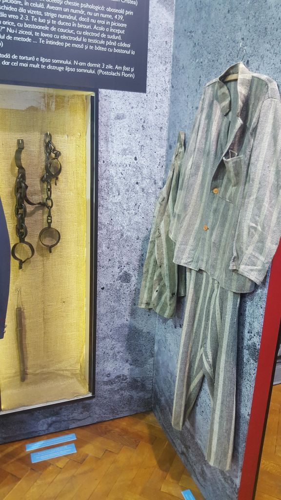 콘크리트와 같은 배경을 가진 줄무늬 유니폼과 녹슨 족쇄가 유리 뒤에 전시되어 있습니다.