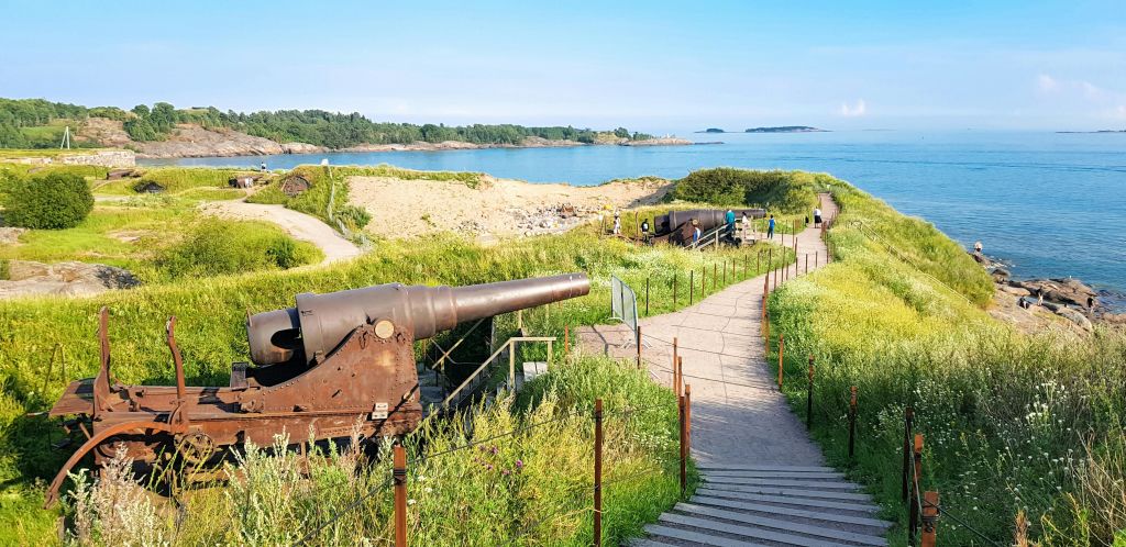 산책로와 푸른 초목이 어우러진 아름다운 해안 풍경이 내려다보이는 녹슨 대포와 소수의 사람들이 걸어다니고 있습니다.