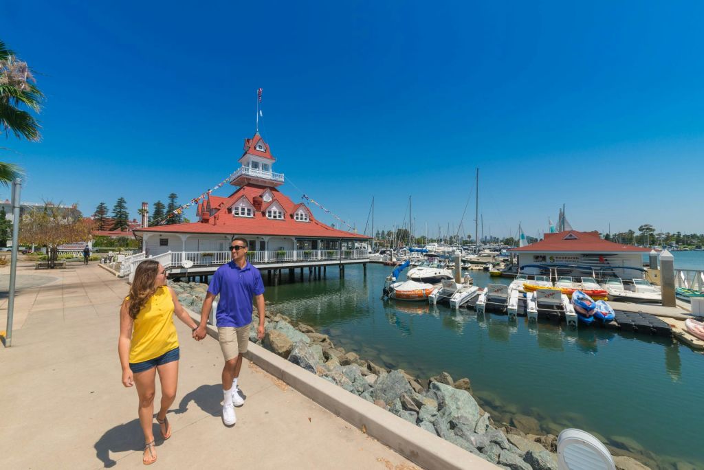 보트를 정박하고 붉은 지붕의 건물을 배경으로 마리나 산책로를 따라 손을 잡고 걷고 있는 커플의 모습입니다.