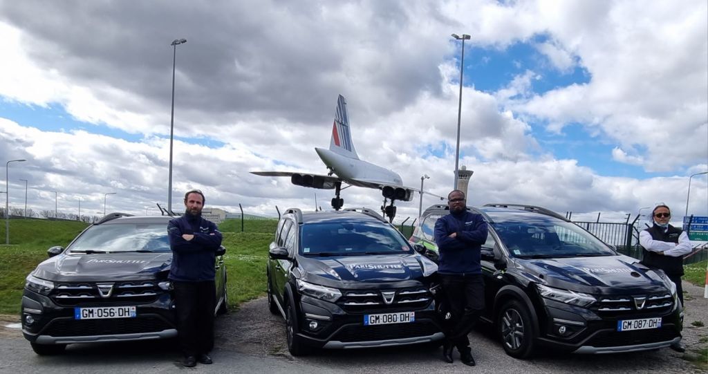한 남성이 프랑스 번호판이 달린 주차된 차량 두 대 사이에 서서 흐린 하늘을 바라보고 있습니다. 비행기가 배경으로 보입니다.