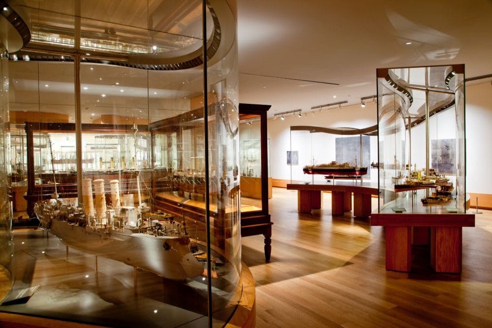나무 바닥과 천장 조명이 있는 유리 케이스에 디테일한 선박 모델이 전시된 박물관 전시관입니다.