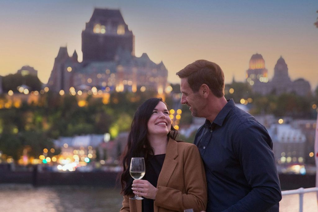 도시 풍경과 조명이 켜진 건물을 배경으로 한 쌍의 커플이 와인 한 잔을 들고 서로 미소 짓고 있습니다.