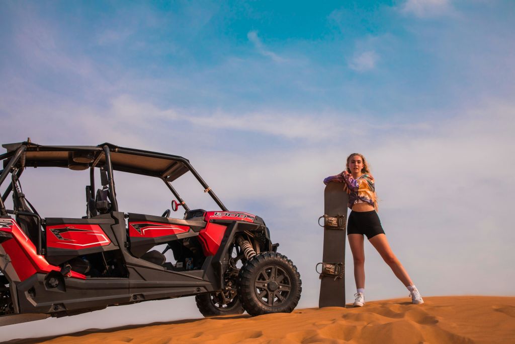 한 여성이 붉은 모래 언덕 버기 옆 사막에 서서 구름이 낀 푸른 하늘 아래 샌드보드를 들고 있습니다.