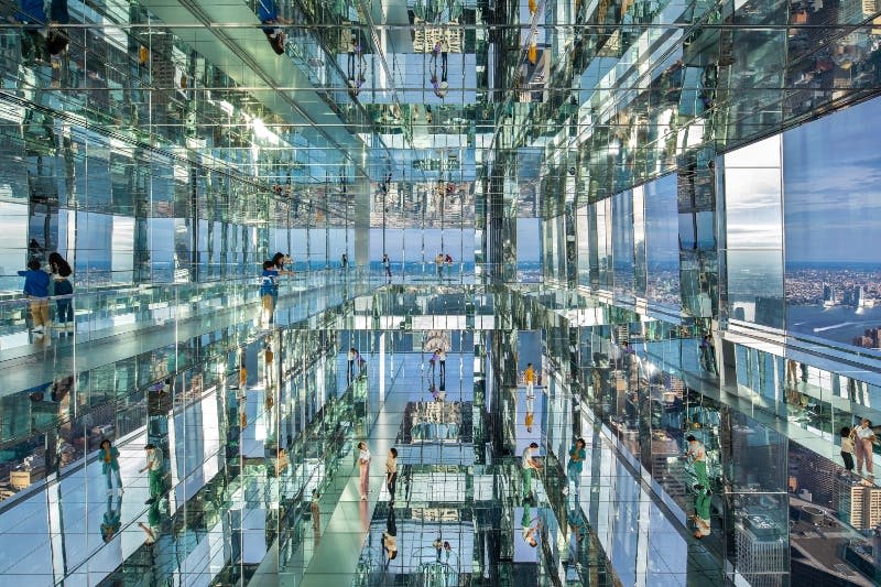 거울로 된 벽과 바닥이 있는 방에서 사람들이 걸어다니며 사진을 찍으며 여러 가지 모습을 연출합니다. 창문을 통해 도시 풍경이 보입니다.