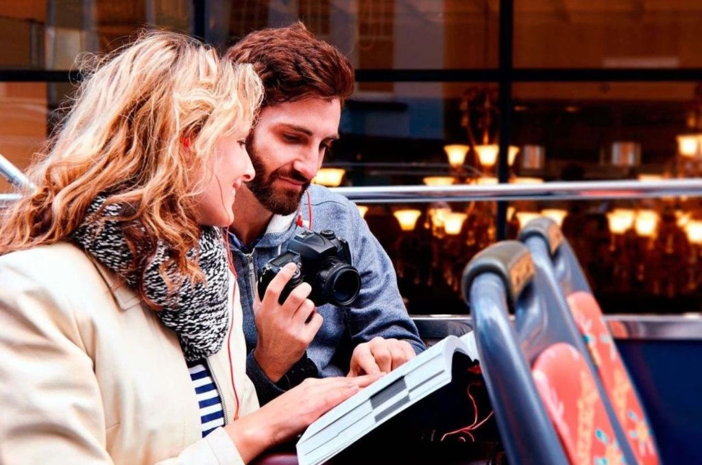 두 사람이 함께 책을 보고 있고, 한 사람은 카메라를 들고 있습니다. 야외에서 반사적인 분위기를 느낄 수 있습니다.