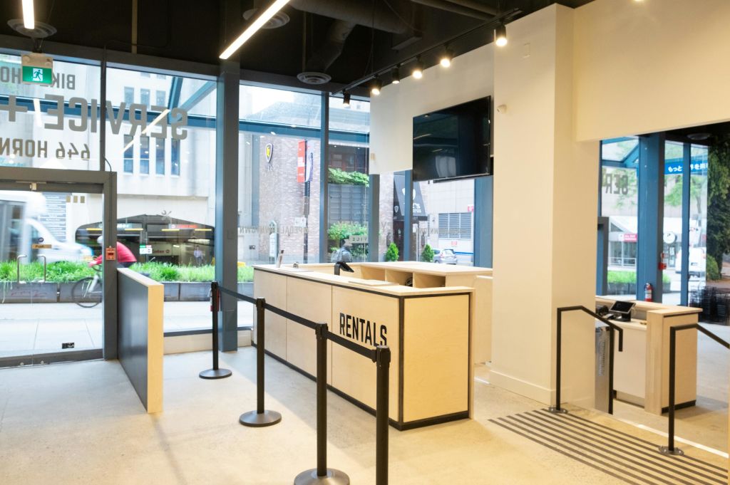 조명이 밝은 현대적인 공간에 '대여' 표지판, 대기열 장벽, 벽걸이 스크린이 있는 실내 대여 카운터.