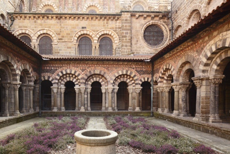 석조 아치와 기둥이 있는 중세 마당으로 중앙의 석조 우물과 전경에 꽃이 피는 식물이 있습니다.