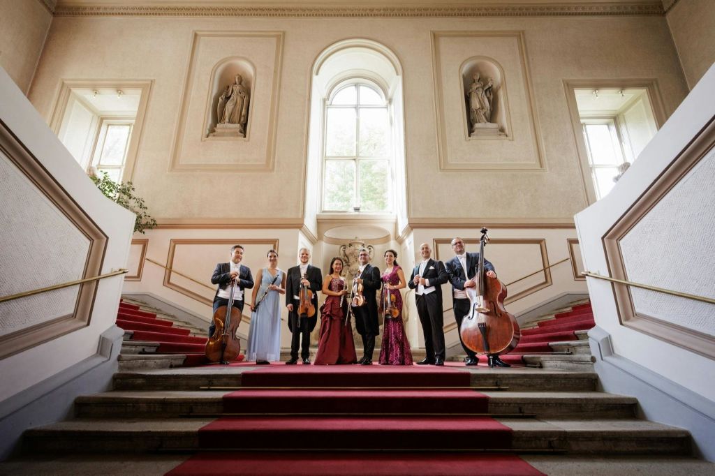 커다란 창문과 조각상이 놓인 그랜드 홀의 레드카펫이 깔린 계단에 현악기를 든 뮤지션들이 서 있습니다.