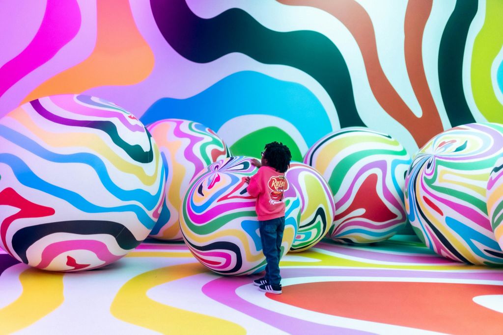 분홍색 재킷을 입은 어린이가 벽과 바닥에 물결무늬가 있는 활기찬 방에서 커다란 다채로운 줄무늬 공과 상호작용합니다.