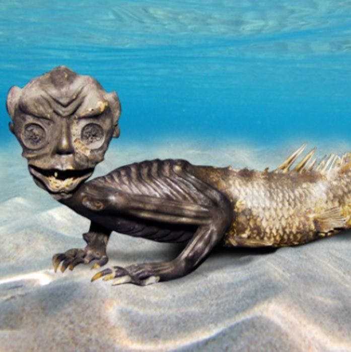 인간형 머리와 물고기 같은 몸을 가진 수상 생물체가 맑은 물 아래 모래 바다 바닥에 놓여 있습니다.