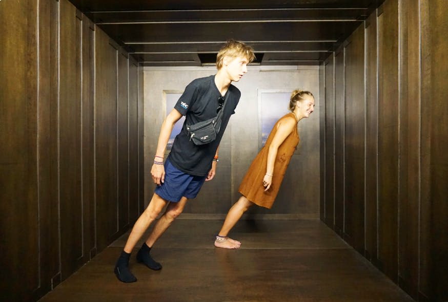 나무 벽과 천장이 있는 기울어진 방에서 두 사람이 부자연스럽게 몸을 앞으로 기울여 착시 현상을 일으킵니다.