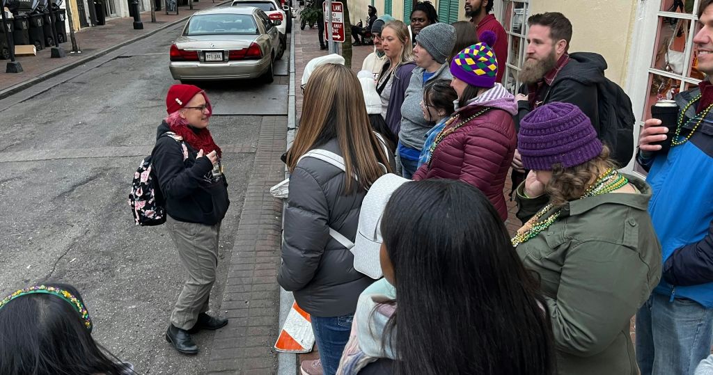 다채로운 색상의 모자와 마르디 그라 (Mardi Gras) 구슬을 쓰고 거리 행사에 참여하는 사람들이 야외에 서 있습니다.