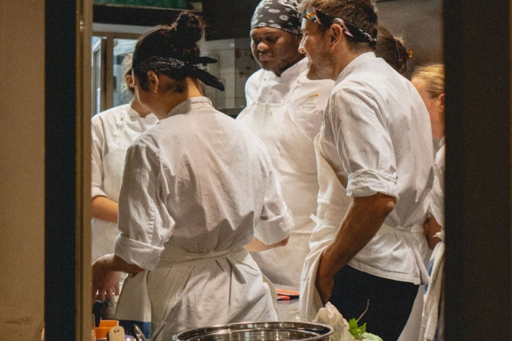 흰색 유니폼을 입은 여러 셰프가 바쁘고 조명이 밝은 주방에서 대화를 나누고 함께 일합니다.