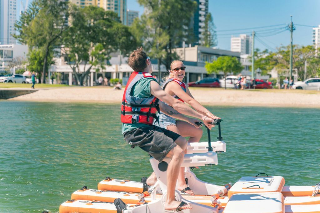 해변 근처에서 선박에 탑승한 두 사람이 구명조끼를 입고 웃고 있으며, 나무와 건물이 배경으로 보입니다.