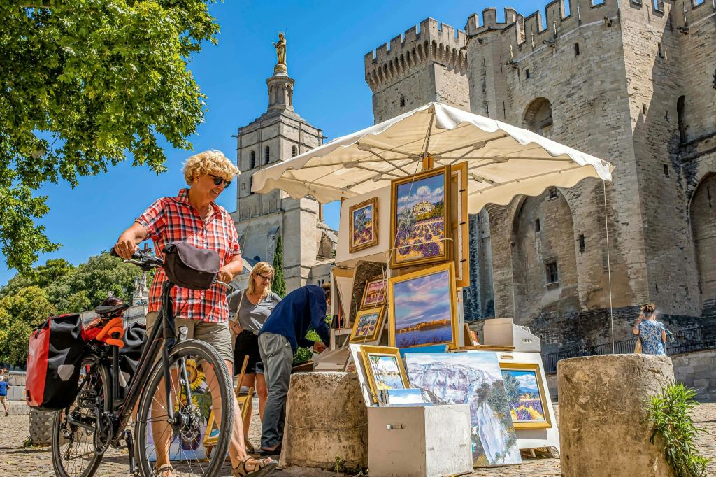 햇살이 내리쬐는 곳에서 탑과 동상이 있는 유서 깊은 석조 건물 근처의 야외 아트 노점을 둘러보는 사람들.