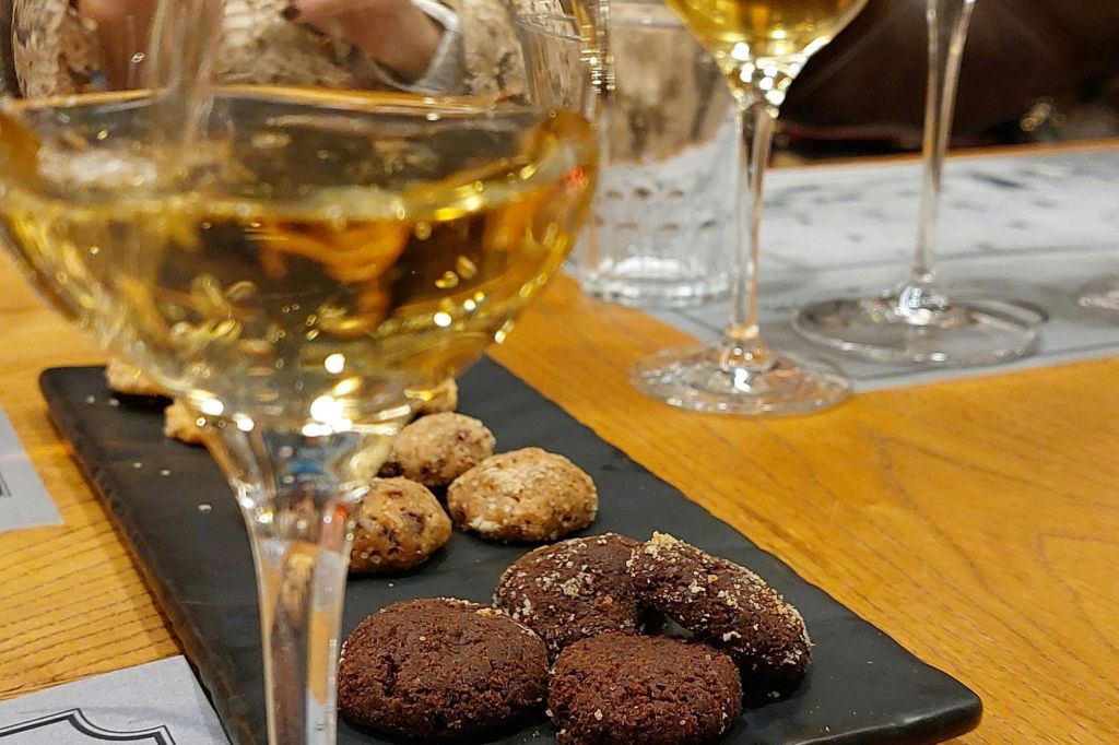 흰색 와인 잔과 검은색 트레이에 모듬 쿠키가 놓여 있으며, 나무 테이블에 사람이 배경으로 놓여 있습니다.