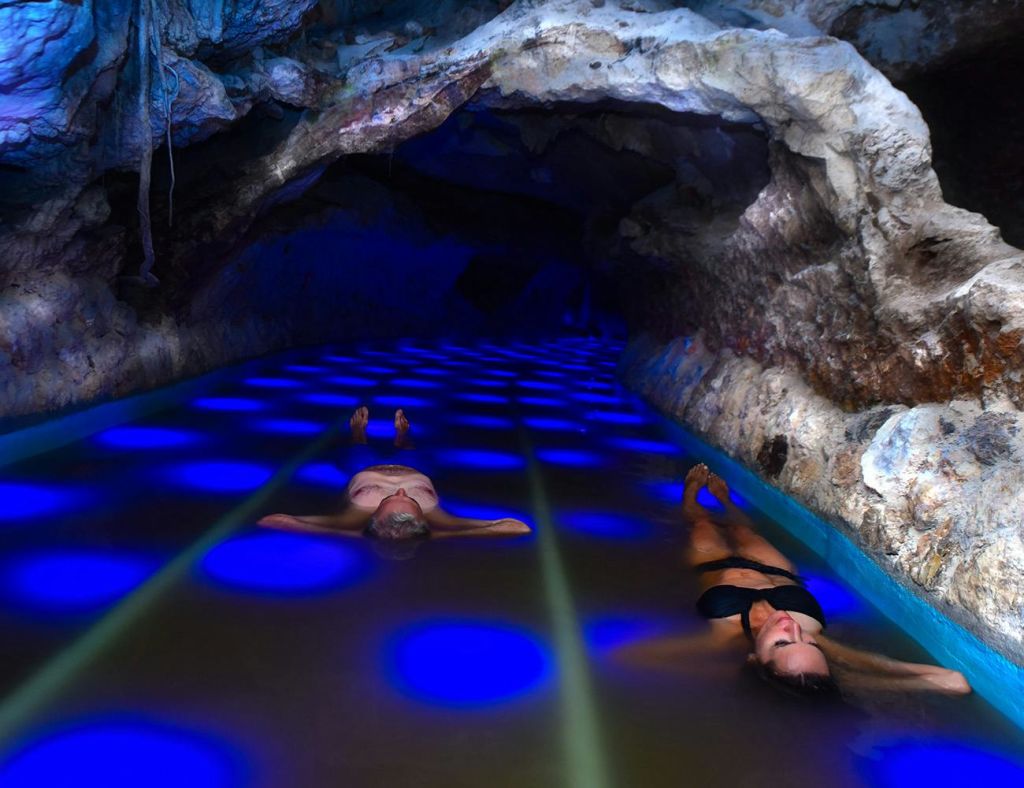 바위 벽으로 둘러싸인 푸른 불빛이 비추는 동굴 수영장에서 두 사람이 등을 맞대고 떠다니고 있습니다.