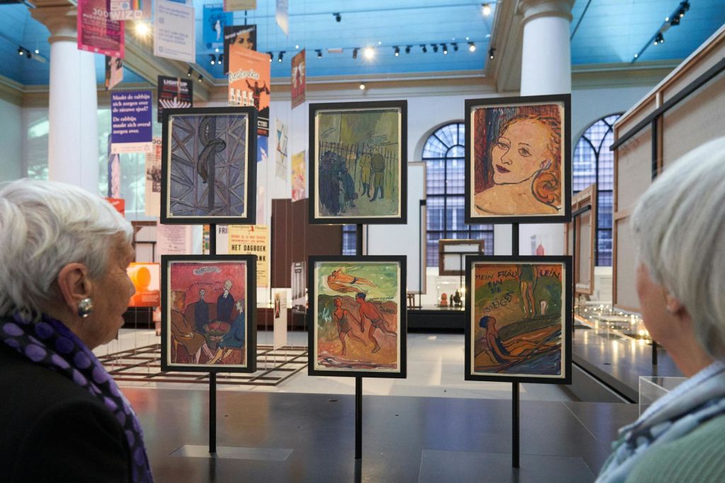 높은 창문과 기둥을 배경으로 네 개의 다채로운 그림이 놓인 박물관에서 액자에 담긴 작품을 감상하는 사람.