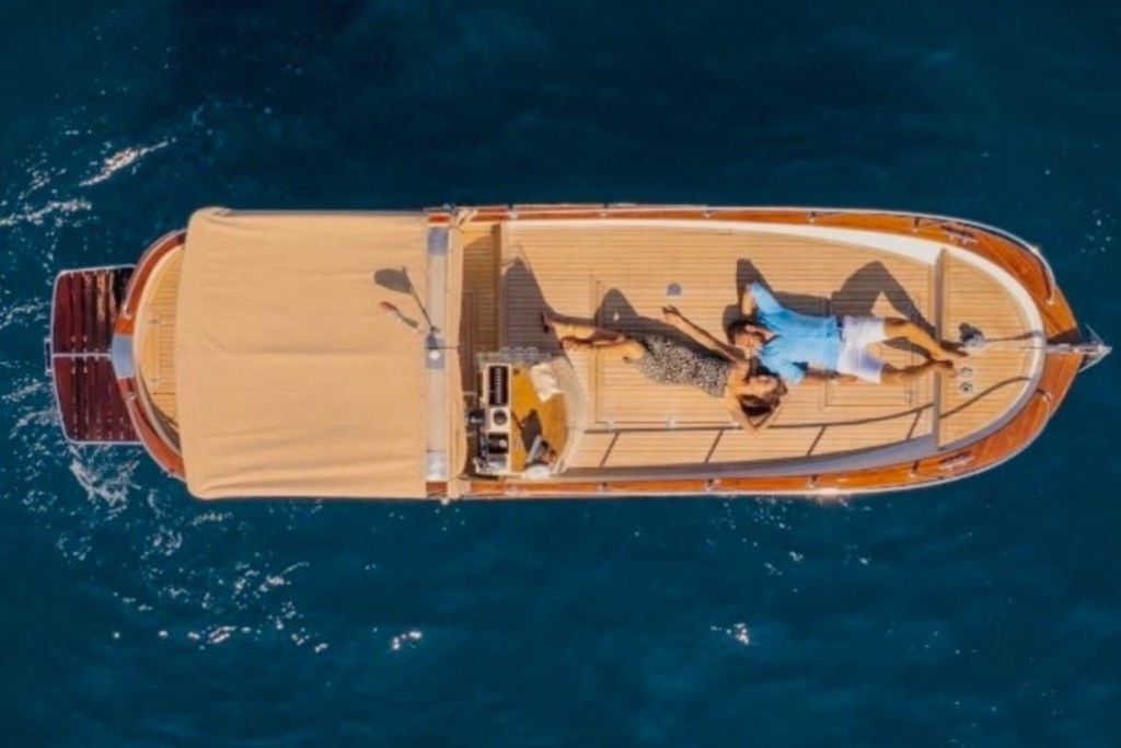 배의 일부를 덮고 있는 베이지색 캐노피로 푸른 물 위의 배 갑판에서 휴식을 취하고 있는 두 사람의 공중 전경.