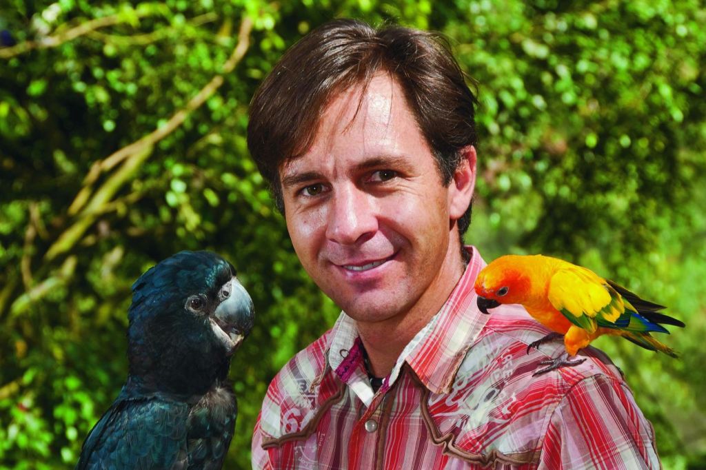 왼쪽에는 검은 앵무새, 오른쪽 어깨에는 노란색 주황색 앵무새를 들고 야외에 서 있는 남성.
