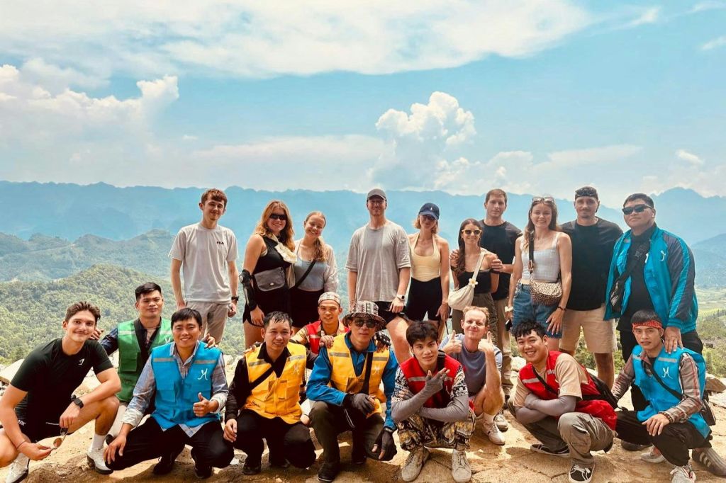 푸르른 언덕과 부분적으로 흐린 하늘을 배경으로 산 전망대에서 포즈를 취하는 13명의 사람들.