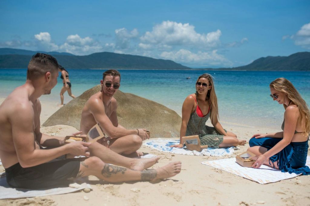 아름다운 바다와 산의 풍경을 배경으로 음식 컨테이너가 놓인 해변에 앉아 미소 짓고 대화하는 세 사람.