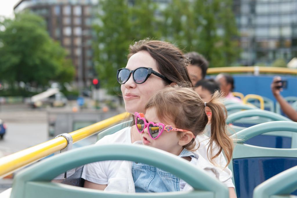 어두운 선글라스를 착용한 여성과 분홍색 하트 모양의 선글라스를 착용한 어린이가 탁 트인 버스를 타고 있으며, 배경에는 흐릿한 나무와 건물이 있습니다.