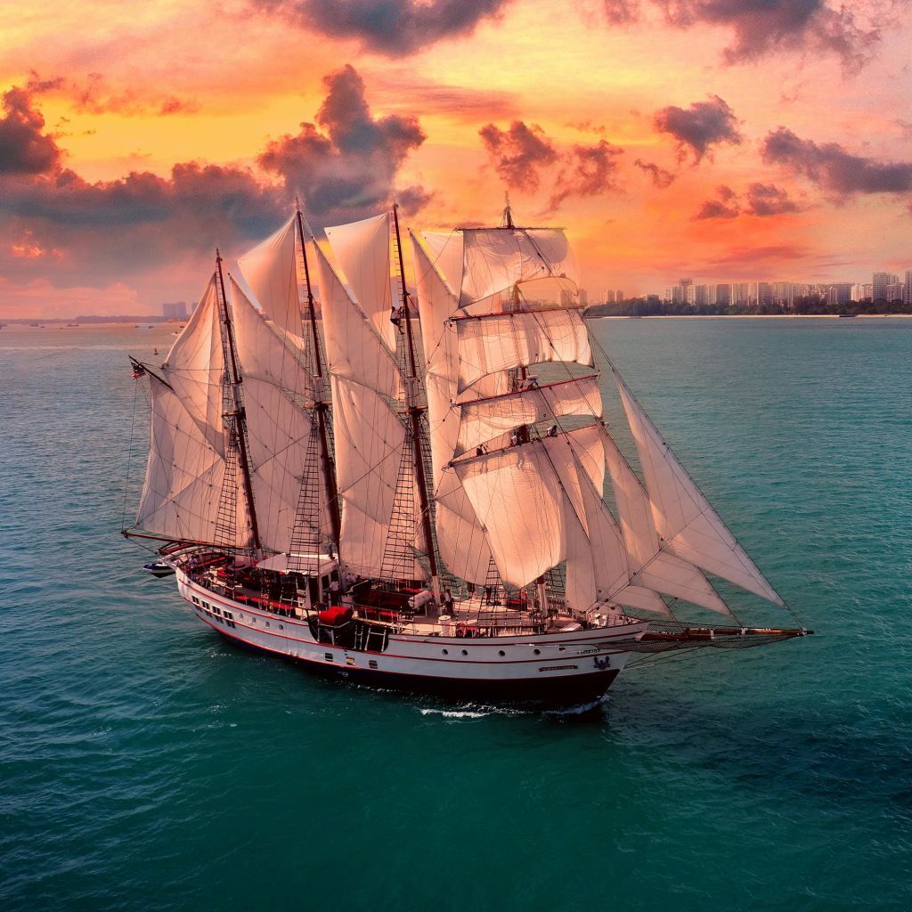 여러 개의 돛이 달린 대형 범선이 일몰에 바다를 항해하고 있으며, 수평선에 멀리 떨어진 도시의 스카이라인이 보입니다.